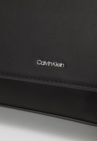 Juodas dirbtinės odos krepšys, turintis tekstūruotą paviršių, elegantišką dizainą ir sidabrinę "Calvin Klein" logotipo detalę priekyje.