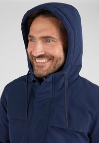 Giacca con cappuccio blu navy con chiusura a zip, caratterizzata da una texture liscia, cordini regolabili e una tasca frontale.