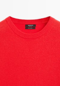 Czerwony sweter z okrągłym dekoltem z kaszmiru z prążkowanym wykończeniem dekoltu oraz metką pokazującą szczegóły marki i materiału.