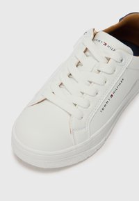 Weißer synthetischer Sneaker mit strukturierten Oberflächen, runder Zehenpartie und Schnürdesign. Mit schwarzer und roter Markenkennzeichnung an der Seite und einem kontrastierenden Innenraum.
