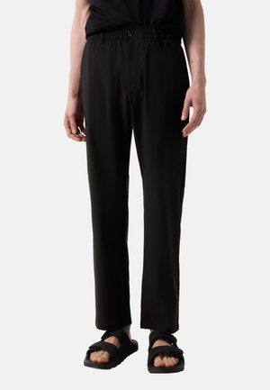 Osoba nosząca czarne spodnie o prostych nogawkach z elastycznym pasem i zapięciem na guzik, w połączeniu z czarnymi sandałami na otwarte palce na białym tle.