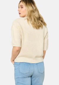 Maglione beige lavorato a maglia con maniche corte, tessuto testurizzato e vestibilità morbida, abbinato a jeans in denim blu chiaro.