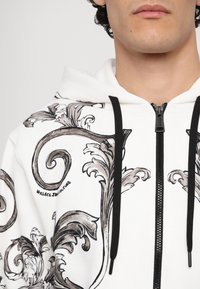 Felpa bianca con intricati motivi a spirale grigi e neri, dotata di zip e lacci neri. "Versace Jeancouture" stampato sul design.