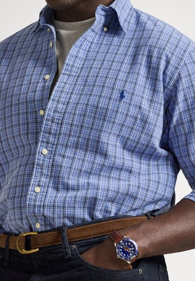 Modra karo srajca s širšimi belimi gumbi, izdelana iz lahkega blaga, združena z rjavim pasom in temnimi kavbojkami. Nosi uro z modrim številčnico.