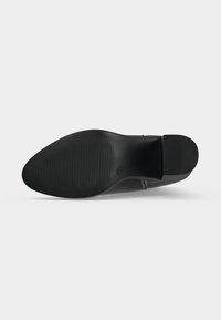 Semelle noire d'une chaussure à bout pointu avec motif texturé antidérapant et petit talon carré sur fond blanc uni.