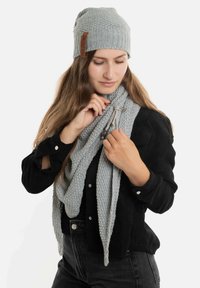 Grijze gebreide muts met een leren label, gecombineerd met een lichtgrijze sjaal met een gemêleerd breiwerk. Model draagt een zwarte blouse en zwarte jeans.