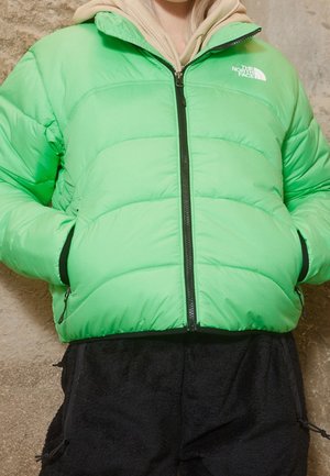 Personne portant une doudoune North Face vert vif par-dessus un sweat à capuche beige, les mains dans les poches de la doudoune, debout contre un mur en pierre.