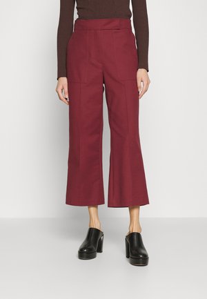 Pantalon large cropped bordeaux associé à des mules à talons hauts noires et un haut marron à manches longues côtelé sur une personne debout.