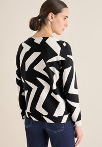 Schwarzer und weißer gemusterter Pullover mit Zickzackdesign, strukturiertem Stoff, Rundhalsausschnitt und langen Ärmeln, kombiniert mit dunkelblauen Jeans.