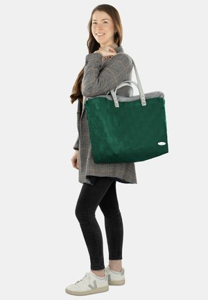 Grüne geflochtene Tragetasche mit grauen Akzenten, ausgestattet mit zwei Tragriemen und einem geräumigen Innenraum. Das Modell trägt einen karierten Mantel und Jeans.