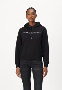 Haut à capuche noir en mélange de coton, avec une poche kangourou à l'avant, un capuchon avec cordon de serrage et un texte logo blanc sur la poitrine. Associé à un jean foncé.