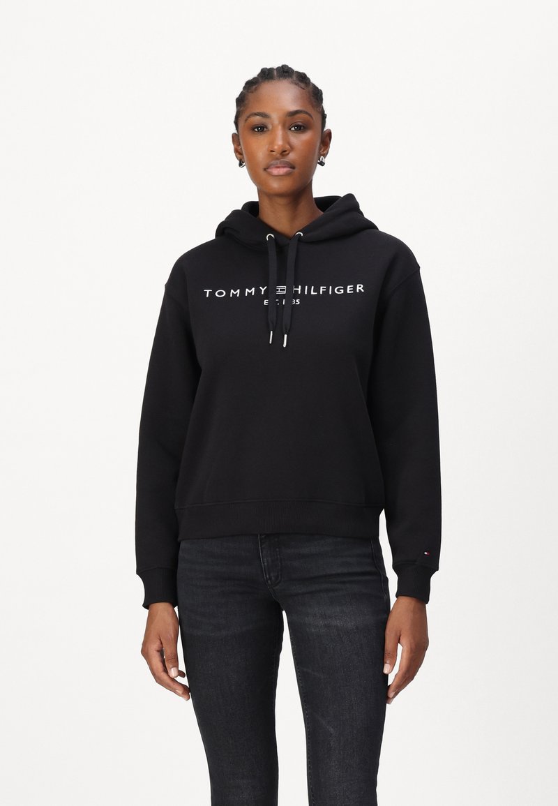 Haut à capuche noir en mélange de coton, avec une poche kangourou à l'avant, un capuchon avec cordon de serrage et un texte logo blanc sur la poitrine. Associé à un jean foncé.