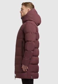 Maroon puffer jacket with a high collar and large hood. Features a zippered side pocket and a textured, quilted design.