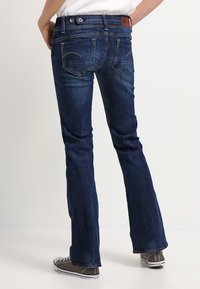 Dark blue denim jeans with a flared leg, featuring twin back pockets, contrasting stitching, and a branded leather patch on the waistband.