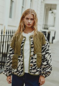 Oliivroheline tikitud jakk mustade ja valgete mustritega fliisseelikutega, millel on kaks eesmist taskut ja puidust nuppe.