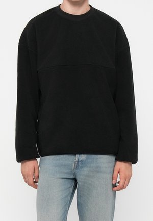 Homme portant un pull polaire noir uni à manches longues et un jean bleu clair, debout devant un fond blanc uni.