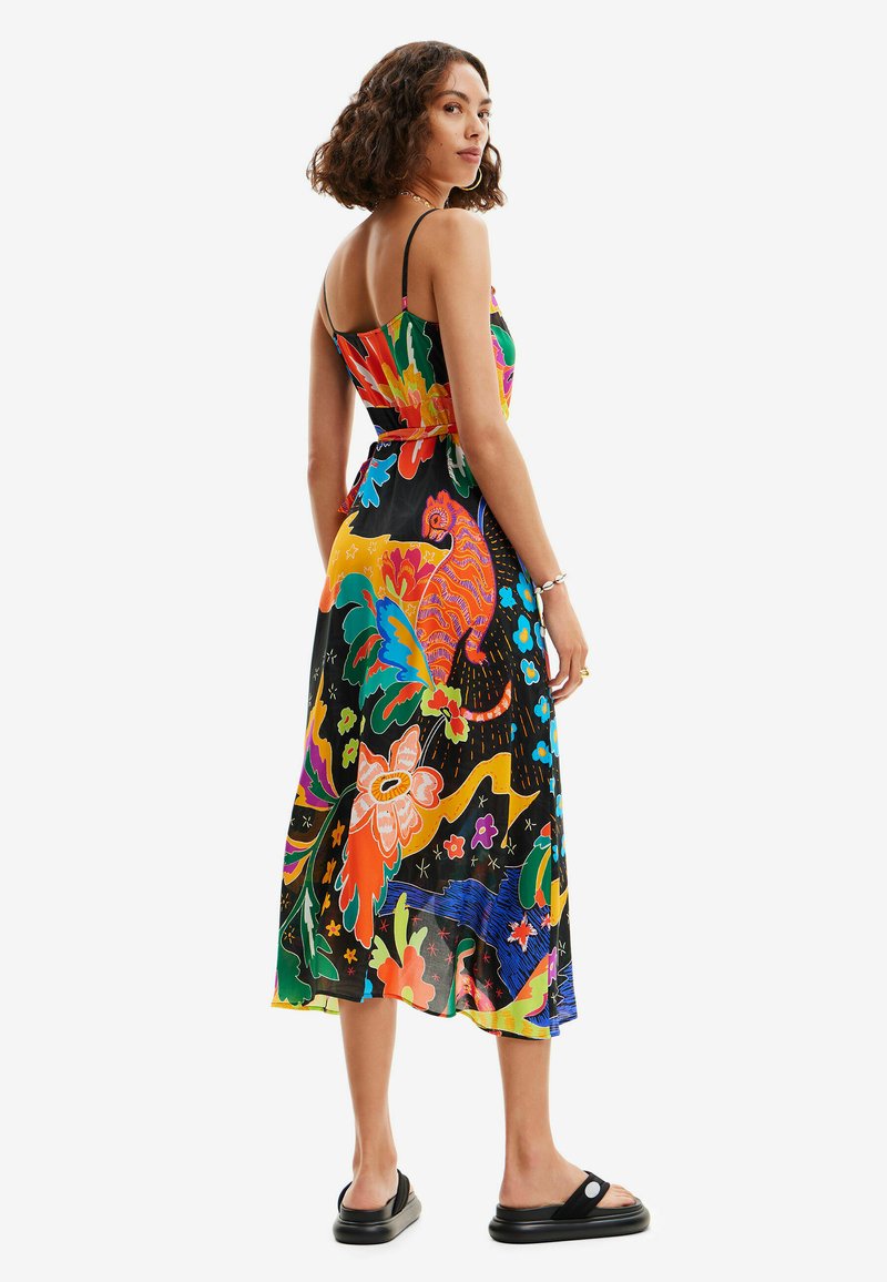 Eine Frau trägt ein farbenfrohes Midi-Kleid mit Blumen- und Tiermuster sowie dünnen Trägern. Sie steht in schwarzen Plateausandalen vor einem weißen Hintergrund.