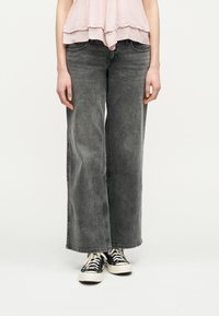 Άτομο που φοράει ξεβαμμένα μαύρα τζιν με φαρδιά μπατζάκια, ασπρόμαυρα αθλητικά παπούτσια και απαλό ροζ τοπ με βολάν, στέκεται μπροστά σε απλό φόντο.