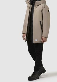 Homme portant un manteau d'hiver beige à capuche avec fermeture éclair noire et poches, pantalon cargo noir et bottes noires à lacets sur un fond clair.