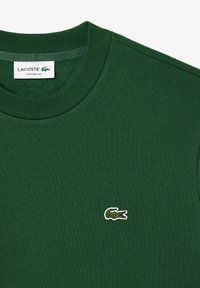 Camiseta de algodón verde con cuello redondo y mangas cortas. Presenta un pequeño logo de cocodrilo bordado en blanco y rojo en el lado izquierdo del pecho.