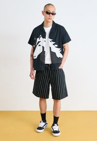 Black short-sleeve shirt with a white horse graphic, paired with pinstriped shorts. Accessories include sunglasses and black sneakers.