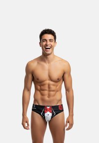 Slip de bain noir avec un design de smoking comportant une chemise blanche et un nœud papillon rouge, accentué par un graphique de rose rouge sur le côté gauche.