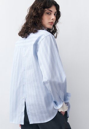 Kobieta o kręconych, ciemnych włosach, ubrana w jasnoniebieską i białą koszulę z pionowymi paskami zapinaną na guziki z tyłu oraz ciemne spodnie, spogląda przez ramię.