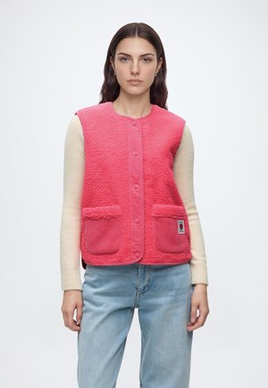Frau trägt eine rosa strukturierte ärmellose Weste über einem cremefarbenen Langarmpullover und hellblauen Jeans, steht vor einem schlichten weißen Hintergrund.