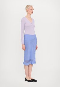 Lila langemouwen top met knopendetail, gecombineerd met een lichtblauwe rok met een gerimpelde zoom en transparante kanten accenten. Zwarte platte schoenen.
