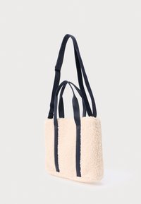 Kreemikas shearling-kott mereväe siniste riidest käepidemetega. Ristkülikukujuline, selgelt nähtava tekstuuriga korpusega ning tasase põhjaga stabiilsuse tagamiseks.