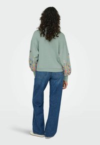 Piparmētru zaļš kapuces krekls ar balonu piedurknēm, uz kura ir ziedu raksts rozā, dzeltenā un zilā krāsā, apvienots ar zilu platu kāju biksēm.