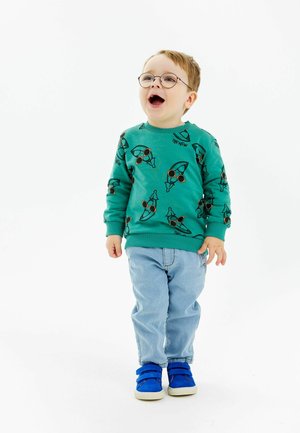 Felpa verde con occhiali illustrati e un design giocoso, abbinata a jeans azzurri e sneakers blu con velcro.