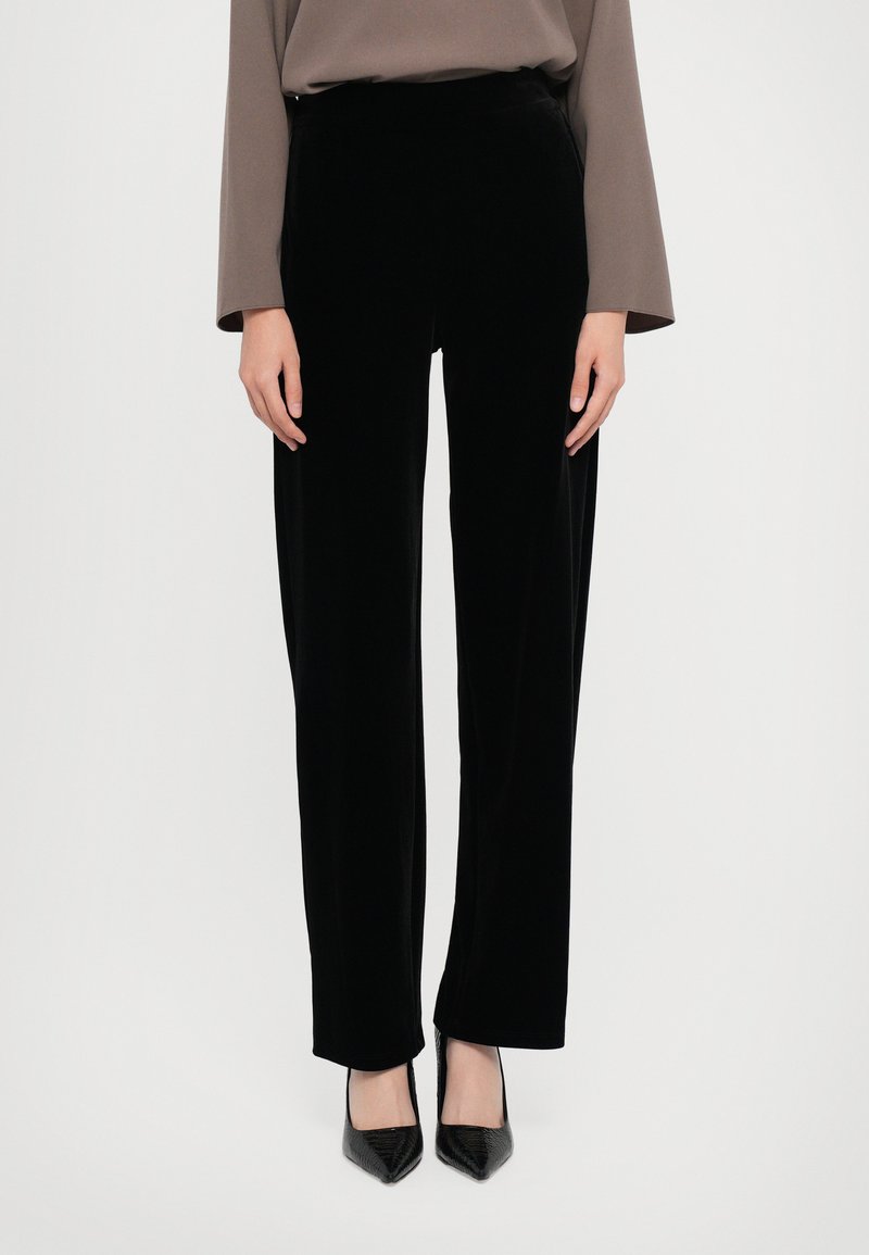 Pantalons en velours noir avec une coupe droite, dotés d'une taille haute et d'une texture lisse, associés à des talons aiguilles noirs.