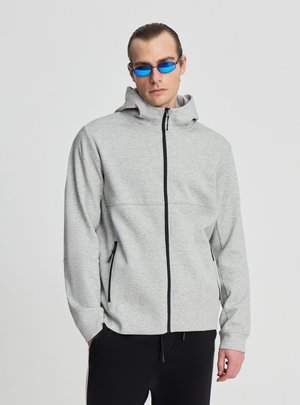 Uomo che indossa una felpa con cappuccio grigio chiaro con zip nera, pantaloni neri e occhiali da sole stretti con lenti leggermente azzurre su sfondo semplice.
