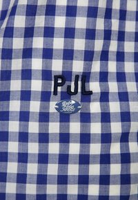 Niebiesko-biała tkanina w kratę z widocznymi wyszywanymi inicjałami "PJL" w czerni oraz okrągłą metką z napisem "Pepe Jeans 1973 London".