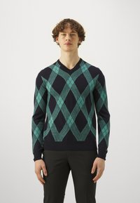 Argyle-mönstrad tröja i marinblått med gröna diamanter. V-ringad design, långärmade, ribbade mudd och nederkant. Stickad textur.