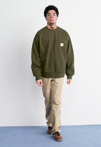 Sudadera verde oliva con detalle de bolsillo frontal, combinada con pantalones beiges y zapatos marrones, sobre un fondo liso.