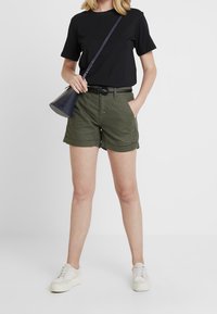 Green shorts with rolled cuffs and belt loops, paired with a black short-sleeve t-shirt and white sneakers, featuring a small black bag.