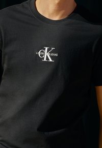 Camiseta de algodón negra con un logo bordado en blanco que dice "Calvin Klein Jeans" y un "CK" estilizado en el centro. Cuello redondo.