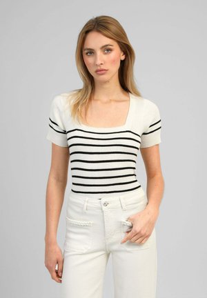 Jeune femme aux cheveux blonds portant un haut blanc à manches courtes avec des rayures horizontales noires et un pantalon blanc taille haute avec un détail corde.