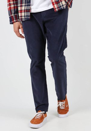 Pantalons en velours côtelé bleu marine avec une coupe droite, associés à des baskets en cuir suédé marron avec des semelles et des lacets blancs. Une chemise à carreaux ajoute un contraste de couleur.