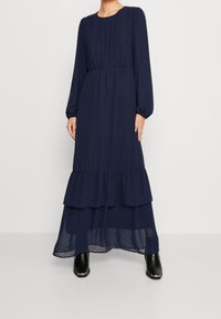 Granatowa sukienka z długim rękawem wykonana z lekkiego materiału, z dopasowaną talią i warstwowym falbanowym dołem, z przezroczystym, kropkowanym wykończeniem u dołu.