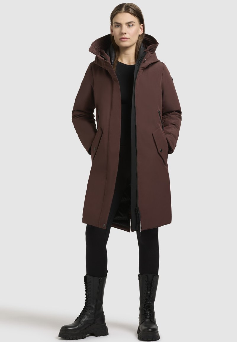 Parka marrone isolata con cappuccio, due tasche frontali e chiusura zip. Presenta una texture liscia e un taglio moderno e allungato.