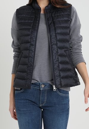 Frau trägt eine schwarze gesteppte ärmellose Weste über einem langärmeligen grauen Pullover und blauen Jeans, wobei die Weste geöffnet ist.