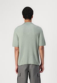 Man i ljusgrön ribbad kortärmad polo tröja och grå cargo-byxor, stående vänd mot en vanlig vit vägg.