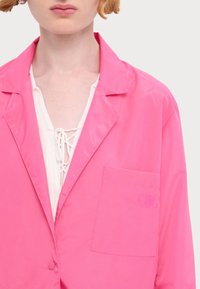 Veste légère rose avec col à revers, fermeture à boutons sur le devant, une poche poitrine unique et un détail brodé subtil sur la poche.