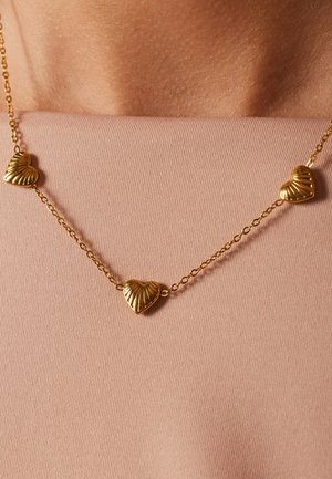 Gouden ketting met drie gegraveerde, hartvormige hangers, gedragen door een persoon met een gladde, lichte huid en een roze stoffen top.