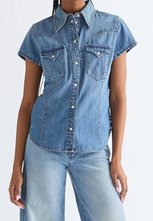 Femme portant une chemise en denim bleu ajustée à manches courtes avec des boutons-pression et deux poches poitrine avant, associée à un jean bleu clair.