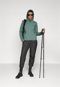 Grønn hettejakke, svarte bukser og turgåerstaver. Jakken har glidelås og to sidelommer, mens buksene har en tapered design.