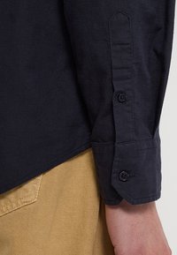 Donkerblauwe shirt met lange mouwen, voorzien van een met knopen sluitende manchette en een lichtelijk gebogen zoom. De stof lijkt textuur en duurzaamheid te hebben.